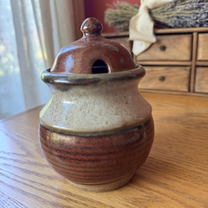 May include: A handcrafted ceramic jar with a lid. The jar is a mix of brown and beige tones, with a textured surface. The lid has a small hole. The jar is sitting on a wooden surface.