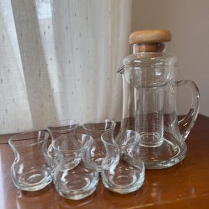 May include: A clear glass tea or coffee press with a wooden lid and handle, accompanied by six small, clear glass cups. The set is arranged on a wooden surface, suggesting a tea or coffee service.