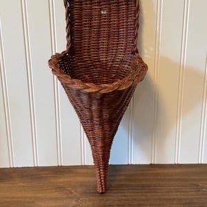 May include: A dark brown woven wicker wall pocket with a cone-shaped base and a flat back panel. The basket features a braided rim detail and a small hole at the top of the back panel for mounting purposes, constructed from natural reed material.