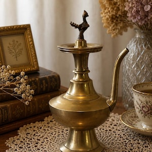 May include: A vintage brass oil lamp with a bird-shaped handle. The lamp has a bulbous base, a slender neck, and a curved spout. It is displayed on a crocheted doily, with a framed picture and a teacup in the background.