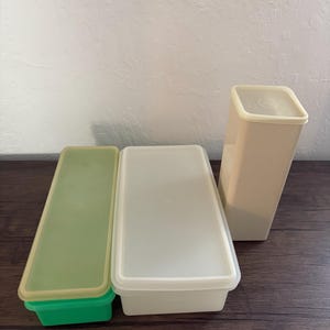 May include: Three plastic storage containers, one green, one white, and one beige. The containers are rectangular and have lids.