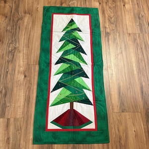 May include: A rectangular quilted table runner featuring a central white panel with a pieced fabric Christmas tree in shades of green. The tree is decorated with gold stitching and sits on a brown trunk and a red base, framed by a green border.