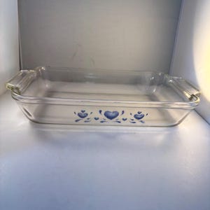 Puede incluir: Una fuente de horno rectangular de vidrio transparente con asas laterales integradas. El exterior frontal presenta un patrón decorativo azul que consiste en tres corazones estilizados con detalles de puntos centrados en el lateral.