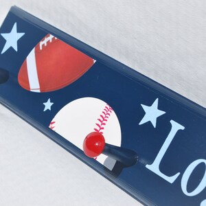 May include: A navy blue wooden wall hook with a baseball, football, and star design. The hook has two pegs with blue and red knobs. The name "Log" is printed in light blue on the right side of the hook.