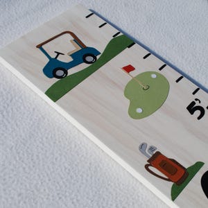 May include: Wooden growth chart featuring golf-themed illustrations. A blue golf cart, a green putting green with a red flag, and a golf bag with clubs are depicted. The chart has black measurement markings and a white background.