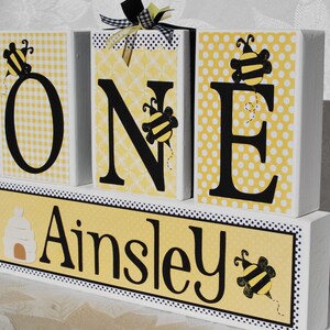 Yellow patterned paper on white wooden blocks spelling out the word 'ONE' in black letters with a bumble bee design on each block. The bottom block is yellow with black polka dots personalized with the name 'Ainsley' with a beehive and a bee design. includes black and white ribbons wrapped around one block. blocks for first birthday.