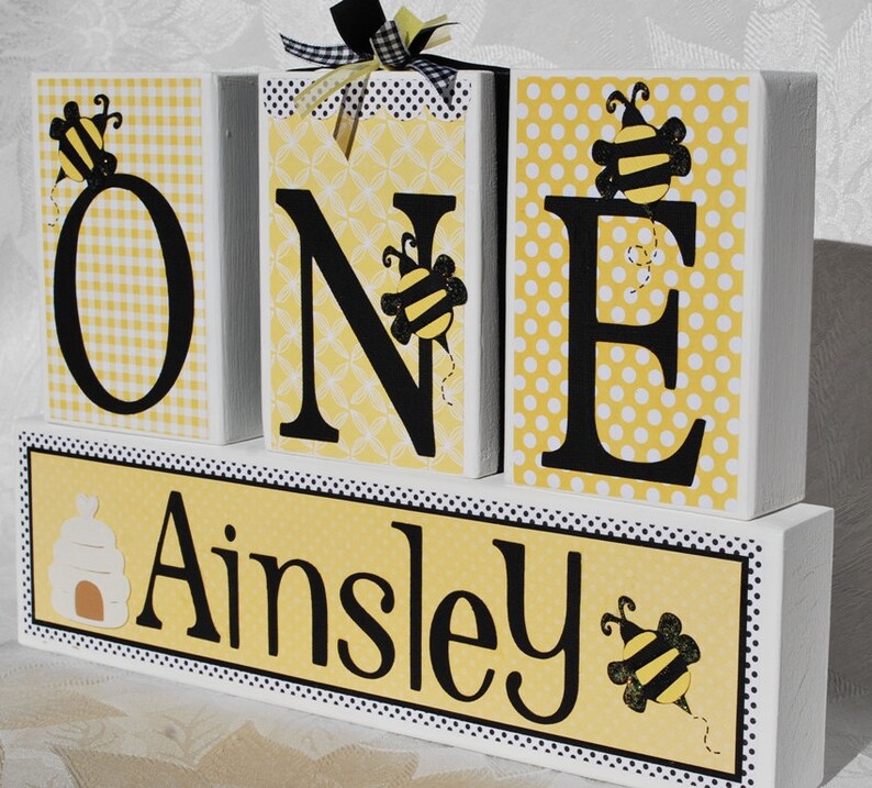Yellow patterned paper on white wooden blocks spelling out the word 'ONE' in black letters with a bumble bee design on each block. The bottom block is yellow with black polka dots personalized with the name 'Ainsley' with a beehive and a bee design. includes black and white ribbons wrapped around one block. blocks for first birthday.