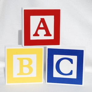 May include: Three colorful wooden alphabet blocks. The top block is red with a white square and the letter "A". The middle block is yellow with a white square and the letter "B". The bottom block is blue with a white square and the letter "C".