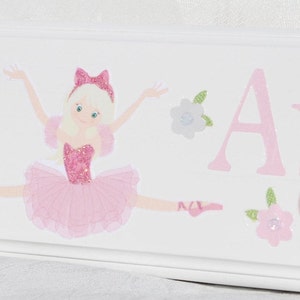 May include: A white wooden wall hook rack featuring a pink glittery ballerina illustration in a split pose, accompanied by small floral accents and the partial name Arde in pink lettering, with a round light pink knob attached to the front.
