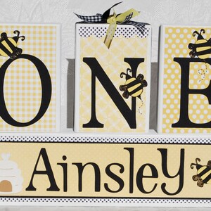 Op de afbeelding: Houten blokken met de tekst "ONE" met bijenaccenten, geplaatst op een basis met de naam "Ainsley". De letters zijn zwart met geel-witte patronen op de achtergrond. De blokken zijn versierd met bijen-thema versieringen en een decoratief lint.