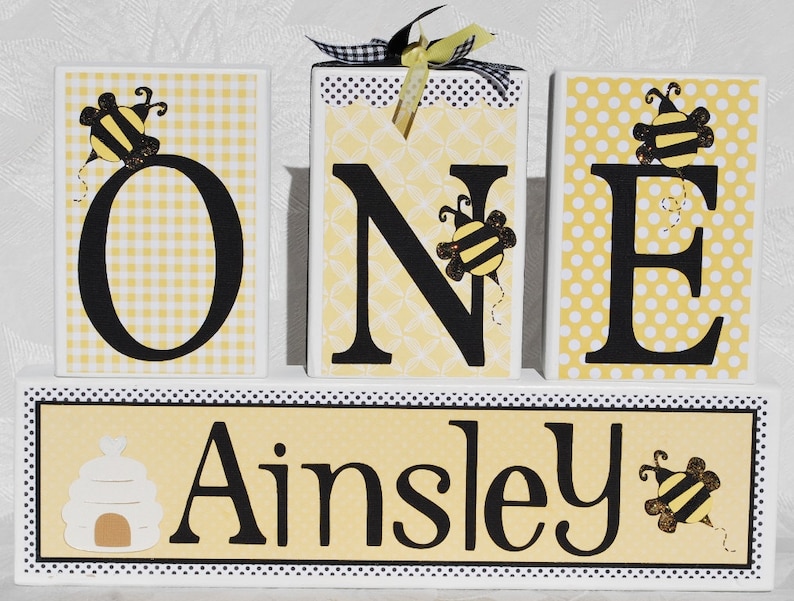 Op de afbeelding: Houten blokken met de tekst "ONE" met bijenaccenten, geplaatst op een basis met de naam "Ainsley". De letters zijn zwart met geel-witte patronen op de achtergrond. De blokken zijn versierd met bijen-thema versieringen en een decoratief lint.