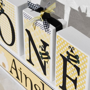 Op de afbeelding: Houten blokken met de tekst "ONE" met decoraties met bijenthema. De letters zijn zwart op gele patronen. Het blok "N" heeft een zwart-wit geruit lint en een geel lint. Het onderste blok zegt "Ainsley".