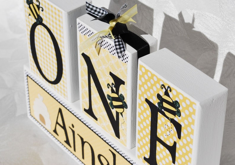 Op de afbeelding: Houten blokken met de tekst "ONE" met decoraties met bijenthema. De letters zijn zwart op gele patronen. Het blok "N" heeft een zwart-wit geruit lint en een geel lint. Het onderste blok zegt "Ainsley".