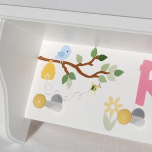 May include: White wooden shelf with a decorative design featuring a blue bird, a beehive, a bee, and the name "Ru" in pink letters. The shelf has two yellow knobs for hanging items.