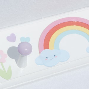 May include: White wooden wall hook with a pastel rainbow, smiling clouds, and a flower design. The hook is lavender and the design is perfect for a child's room. The hook is approximately 2 inches long.