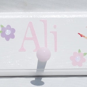 May include: A white wooden wall hook rack featuring illustrations of ballerinas in pink tutus, ballet slippers, and colorful flowers. The name Ali is printed in pink script in the center. The rack includes four round, light pink knobs for hanging.