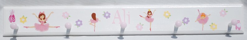 May include: A white wooden wall hook rack featuring illustrations of ballerinas in pink tutus, ballet slippers, and colorful flowers. The name Ali is printed in pink script in the center. The rack includes four round, light pink knobs for hanging.
