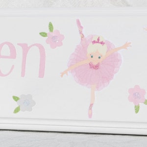 May include: A white wooden wall hook rack featuring the name Arden in pink lettering, decorated with two illustrations of ballerinas in pink tutus and small floral accents, and two round light pink knobs for hanging items.