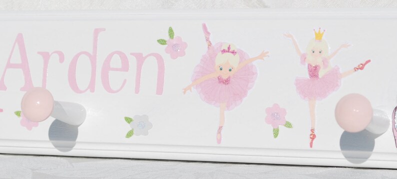 May include: A white wooden wall hook rack featuring the name Arden in pink lettering, decorated with two illustrations of ballerinas in pink tutus and small floral accents, and two round light pink knobs for hanging items.