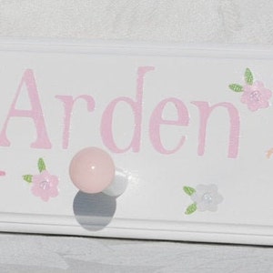 May include: A white wooden wall hook rack featuring the name Arden in pink lettering. The design includes three illustrations of ballerinas in pink tutus and small floral accents. The rack has three round pink knobs for hanging items.