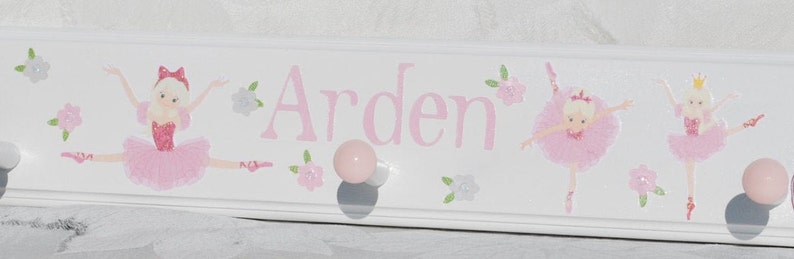 May include: A white wooden wall hook rack featuring the name Arden in pink lettering. The design includes three illustrations of ballerinas in pink tutus and small floral accents. The rack has three round pink knobs for hanging items.
