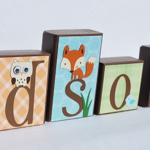 May include: Wooden blocks spelling out a name, featuring an owl, fox, and bird. The blocks have brown sides and tops, with patterned paper fronts in orange, blue, and green. The letters are brown, and the animals are cut out.