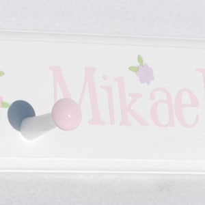 May include: A white rectangular wall-mounted coat rack featuring four hooks with pink ball ends. The surface is decorated with illustrations of ballerinas in pink tutus, ballet slippers, and small flowers, with the name Mikaela printed in pink.