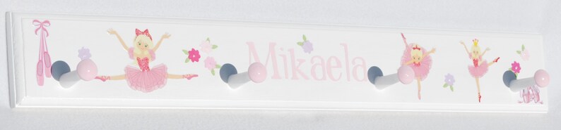 May include: A white rectangular wall-mounted coat rack featuring four hooks with pink ball ends. The surface is decorated with illustrations of ballerinas in pink tutus, ballet slippers, and small flowers, with the name Mikaela printed in pink.