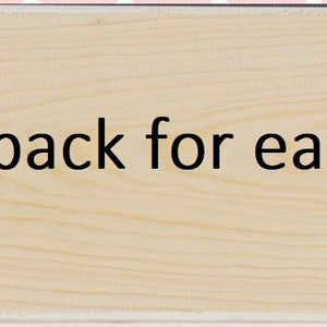 May include: A rectangular light-colored wooden board featuring two vertical keyhole slots carved into the surface. The text "Keyholes in back for easy hanging" is printed in black in the center of the wood grain surface between the two slots.