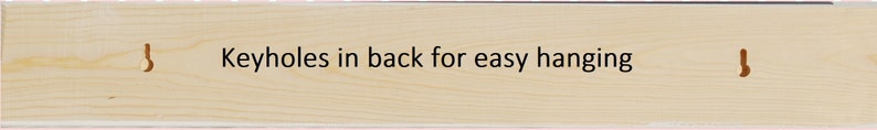 May include: A rectangular light-colored wooden board featuring two vertical keyhole slots carved into the surface. The text "Keyholes in back for easy hanging" is printed in black in the center of the wood grain surface between the two slots.