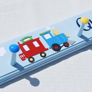 May include: A light blue wooden coat rack with a train design. The train is composed of a red caboose and a blue engine. The name "Quinn" is written in blue script. The rack has two pegs with a blue and yellow top.