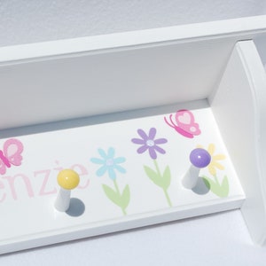 May include: White wooden shelf with two pegs, decorated with a floral and butterfly design. The shelf has a pink butterfly and the name "kenzie" in pink. The pegs are yellow and purple.