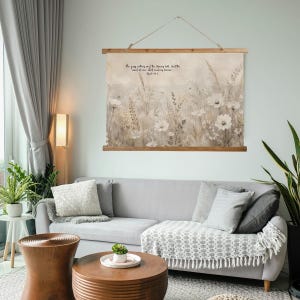 Könnte beinhalten: Ein floraler Wandbehang mit Holzrahmen und Aufhängeschnur. Das Kunstwerk zeigt weiße Blumen und beiges Laub vor neutralem Hintergrund. Text ist in der oberen linken Ecke sichtbar. Der Wandbehang wird über einem grauen Sofa präsentiert.