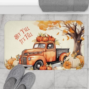 May include: A rectangular bath mat featuring an orange vintage truck loaded with pumpkins. The mat has the text "HEY Y'ALL IT'S FALL" in red. The background includes a tree with orange leaves and pumpkins.