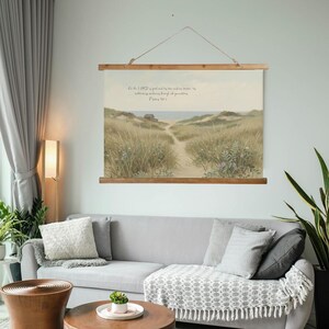 Puede incluir: Un tapiz de pared de lona con una escena de paisaje costero. La obra de arte representa un camino arenoso que conduce al océano, enmarcado por hierbas altas y flores silvestres. El texto en la parte superior dice: "Porque el Señor es bueno y su amor perdura para siempre; su fidelidad por todas las generaciones. Salmo 100:5."