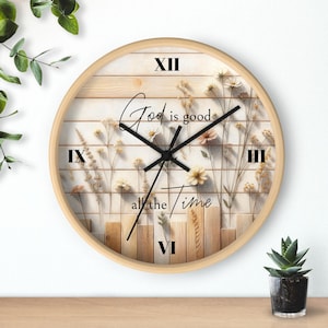 May include: Circular wall clock with a light wood frame. The clock face has a light wood background with floral accents and the text "God is good all the time" in black script. Roman numerals mark the hours.