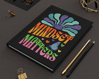Könnte beinhalten: Schwarzes Notizbuch mit den Worten "MINDSET MATTERS" in farbenfroher Retro-Schrift, mit einer Blumen- und Schmetterlingsgrafik. Goldene und schwarze Bleistifte und goldene Büroklammern liegen auf einer dunkelgrauen Oberfläche.