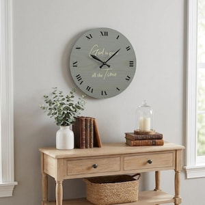 Op de afbeelding: Een ronde, grijze houten wandklok met zwarte Romeinse cijfers en wijzers. De wijzerplaat heeft de tekst "God is good all the time" in een scriptlettertype. De klok hangt boven een houten console.