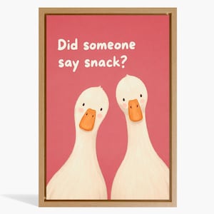 Könnte beinhalten: Ein gerahmter Kunstdruck mit zwei weißen Enten mit orangefarbenen Schnäbeln vor einem einfarbigen rosa Hintergrund. Der Text Did someone say snack? ist in Weiß in der oberen linken Ecke gedruckt. Das Bild ist in einem hellen Holzrahmen.
