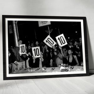 May include: A black and white framed photograph depicting a crowd of people holding up white scorecards with the number 10 printed in bold black ink. The scene captures a lively event with individuals seated at a table, displaying their scores.