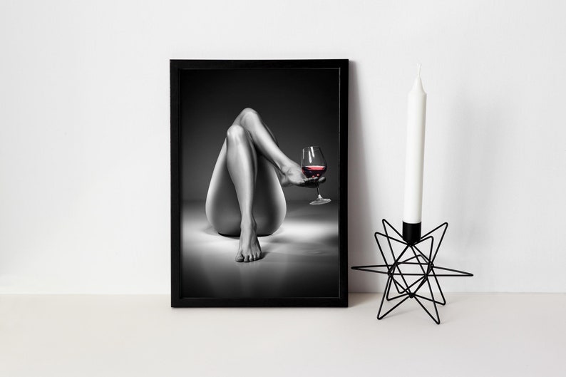 May include: Black and white framed print of a person holding a glass of red wine. A black metal starburst candle holder with a white candle is to the right of the framed print. The image is a sensual, artistic depiction.