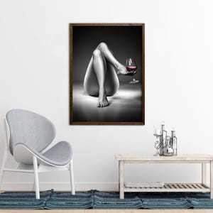 May include: Black and white framed art print featuring a grayscale image of a person's legs and torso, holding a glass of red wine. The artwork is displayed on a white wall, with a gray chair and a wooden table in the foreground.