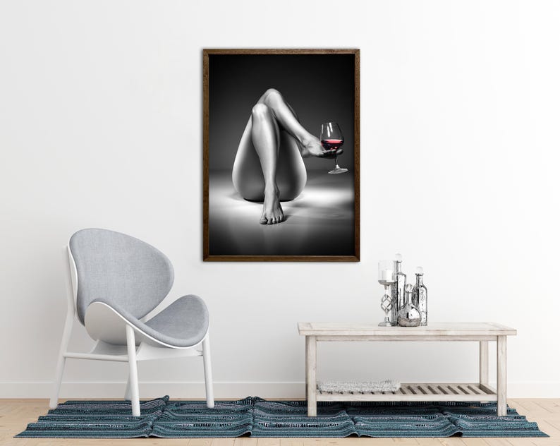 May include: Black and white framed art print featuring a grayscale image of a person's legs and torso, holding a glass of red wine. The artwork is displayed on a white wall, with a gray chair and a wooden table in the foreground.