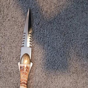 Puede incluir: Un cuchillo decorativo con una hoja plateada y un mango con forma de pez. El mango presenta una parte superior dorada y un material similar al hueso con un patrón marrón. El cuchillo mide unos 25 cm de largo.