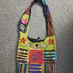 May include: A colorful patchwork shoulder bag with a peace sign and floral embroidery. The bag is made of different fabrics and has a long strap.