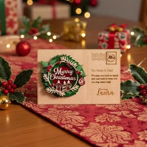 May include: A wooden Christmas card with a wreath design and the words "Merry Christmas." The card includes a message to Mom & Dad and is signed "Loving Laura." The card is on a red floral runner.