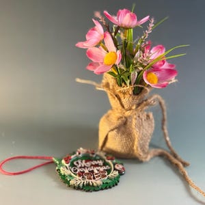 May include: A small burlap sack holds pink flowers with yellow centers and green stems. A Christmas ornament with the words "Merry Christmas" is next to the sack. The ornament has a green wreath design with red accents and a red string.