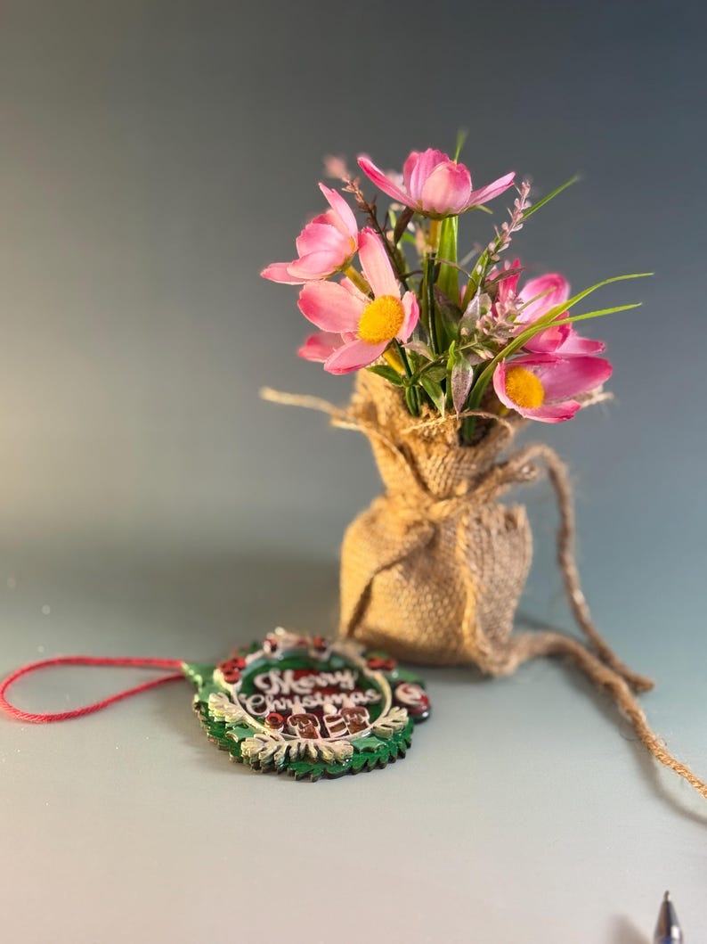 May include: A small burlap sack holds pink flowers with yellow centers and green stems. A Christmas ornament with the words "Merry Christmas" is next to the sack. The ornament has a green wreath design with red accents and a red string.