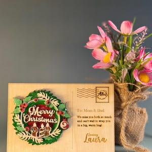 May include: A wooden Christmas postcard with a wreath design and the words "Merry Christmas." The postcard includes a message to "Mom & Dad" and is decorated with a Santa stamp and pink flowers in a burlap vase.