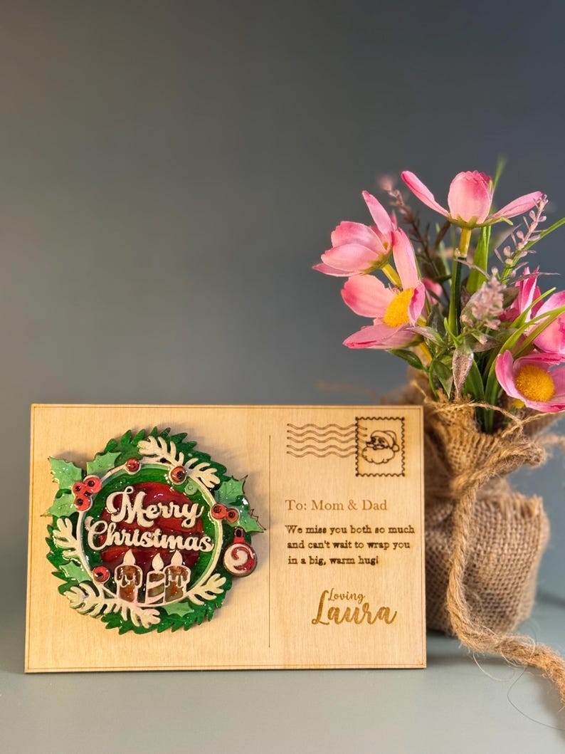 May include: A wooden Christmas postcard with a wreath design and the words "Merry Christmas." The postcard includes a message to "Mom & Dad" and is decorated with a Santa stamp and pink flowers in a burlap vase.
